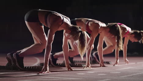 A-row-of-runners-womens-crouch-in-the-starting-position-before-beginning-to-race.-Females-start-with-running-shoes-on-the-stadium-from-the-start-line-in-the-dark-with-spotlights-in-slow-motion.